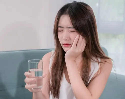Woman holding glass of water, touching cheek in discomfort, suggesting dental pain or sensitivity.
