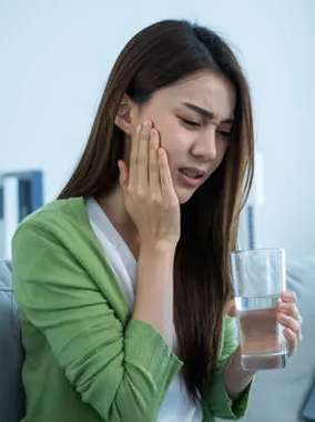 Woman in green shirt holding cheek in pain, holding water glass, indoor setting.