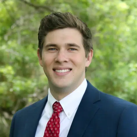 Professional headshot of man in dark blazer and red tie against green foliage background.